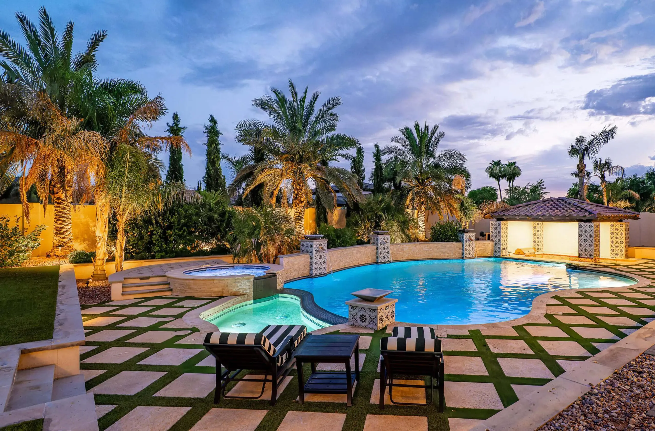 A luxurious backyard in Buckeye, AZ features a large pool with adjacent jacuzzi, surrounded by palm trees and well-manicured shrubs. The patio area includes striped cushioned lounge chairs and a small table. Soft evening lighting illuminates the low-maintenance lawn installed by an expert artificial grass installer.