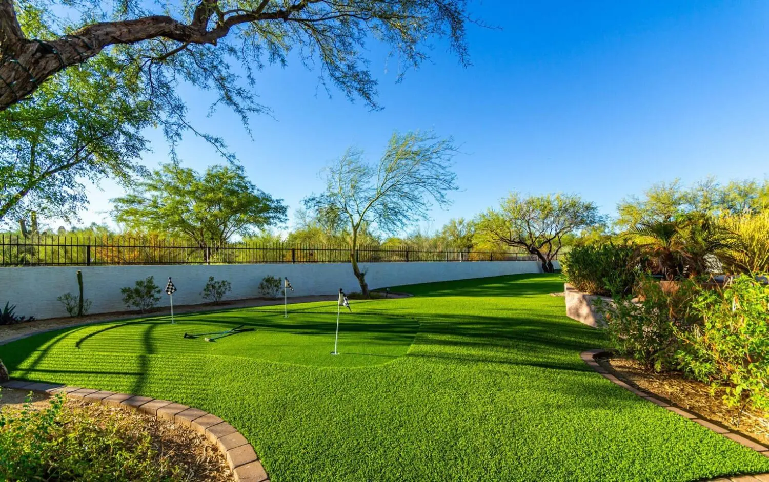 A lush backyard features a well-maintained putting green under a clear blue sky. Surrounded by trees and a white fence, the green—crafted from premium synthetic grass—has multiple small flags marking holes, and a brick pathway outlines its edge. Desert plants are scattered around the yard.