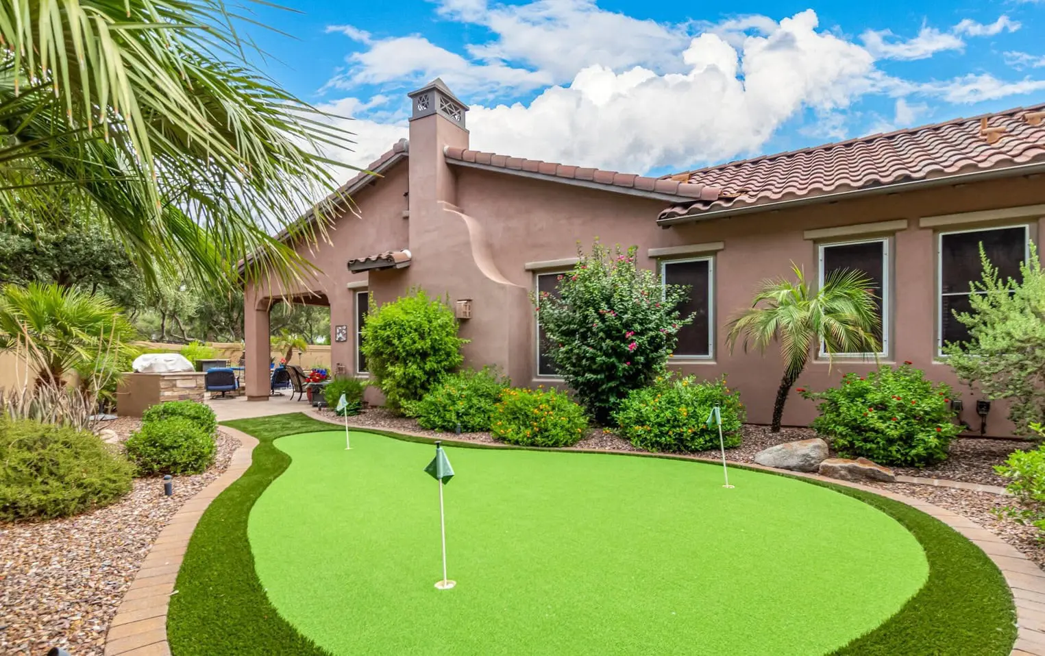 A small, curly-haired dog runs joyfully on a lush green lawn made of artificial grass with its mouth open and ears flapping. The background features various plants, including Cactus Turf, and bushes. The scene is bright and sunny, suggesting a warm day in Buckeye AZ.