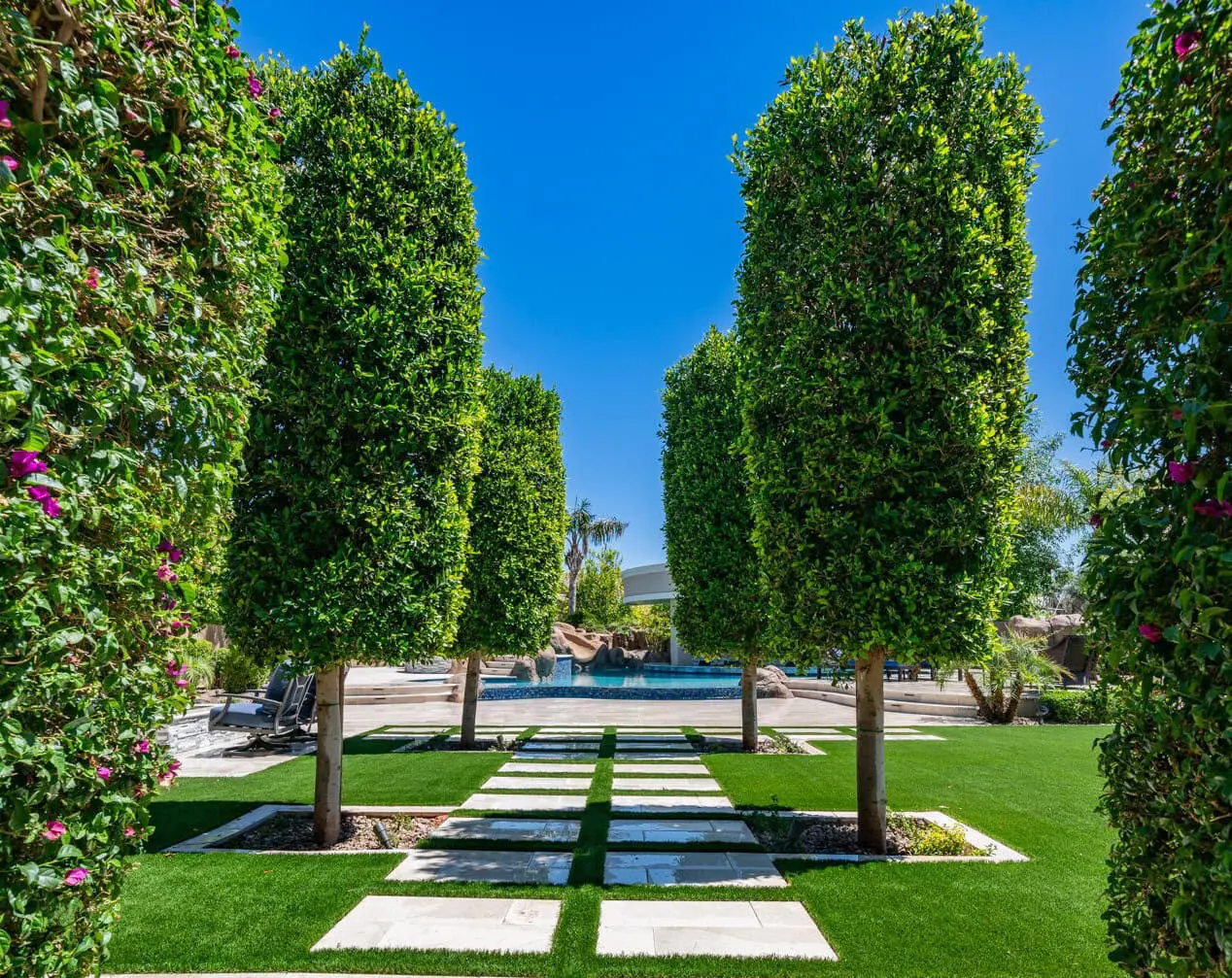 A well-manicured garden with tall, square-shaped trees lining a pathway of rectangular stone slabs. The path leads to a swimming pool surrounded by lush lawns and a bright blue sky, creating an oasis-like retreat. For those interested in achieving this look, contact your local artificial grass installer for a free quote.