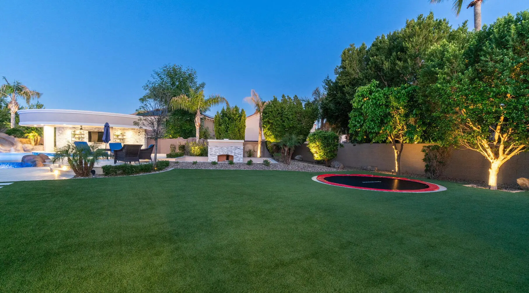 A Buckeye, AZ backyard with a green lawn, an in-ground trampoline, trees and shrubs along the back fence, a patio with outdoor furniture, and a swimming pool with a slide and umbrellas. The sky is clear, the area is well-lit with daylight, and the turf is lush thanks to Cactus Turf.