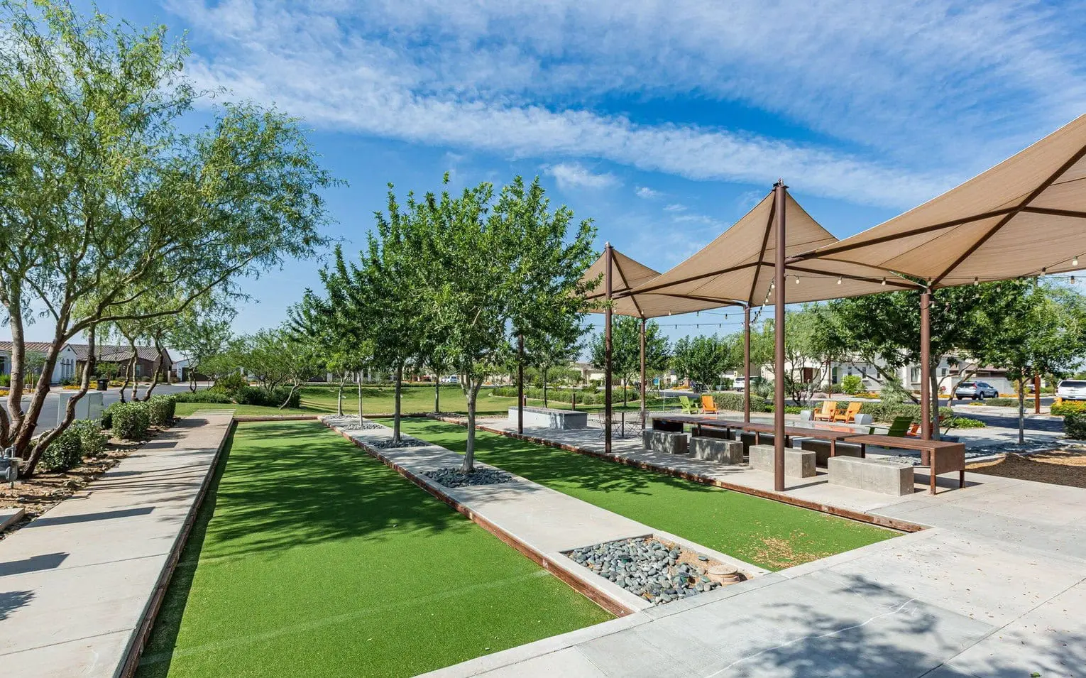 A landscaped park area in Buckeye, AZ features a shaded picnic area with tables and benches under beige triangular canopies. Adjacent is a green lawn strip with pristine cactus turf, bordered by pathways and surrounded by trimmed trees under a bright blue sky. Relaxing seating is visible in the background.