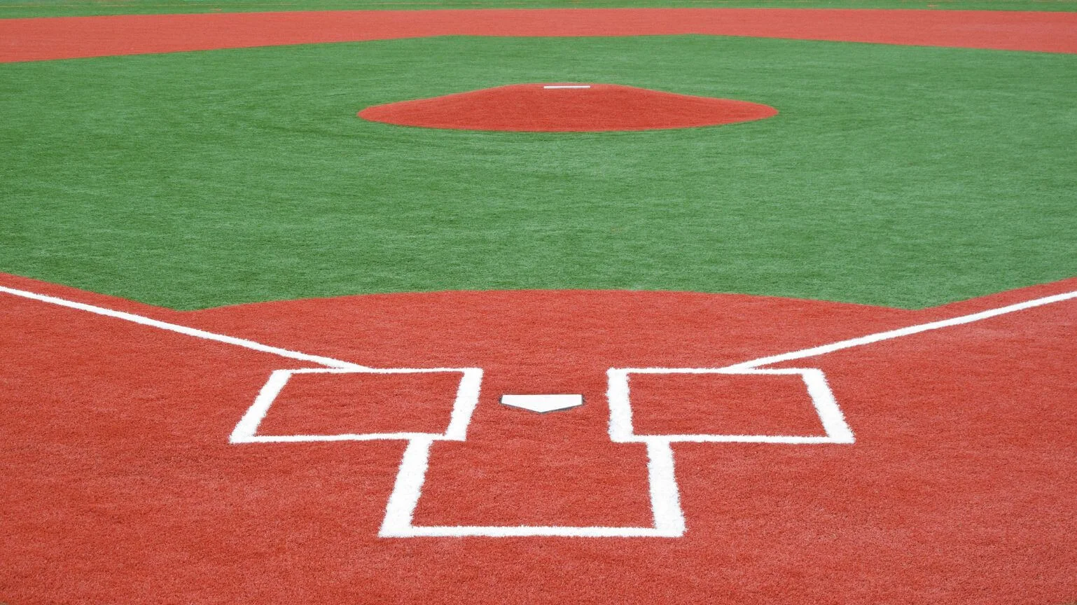 A close-up view of a baseball field's home plate area is seen, featuring the white rectangular batter's boxes outline and the small pentagonal home plate on a red dirt base. Beyond it, the artificial grass turf extends towards the pitcher's mound in the distance, showcasing sports field installations in Buckeye, Arizona.