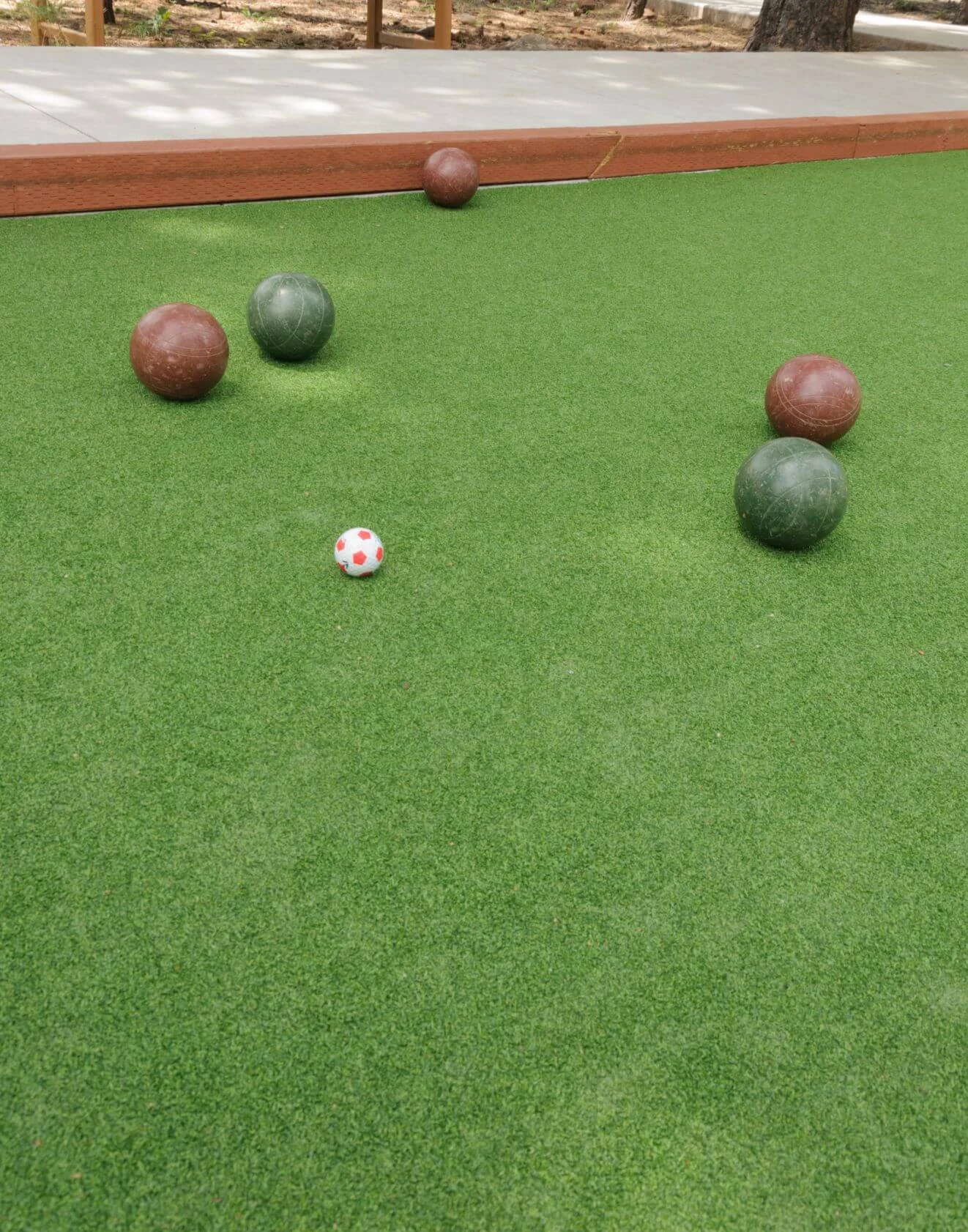 A bocce ball court in Buckeye, Arizona, with four large balls (two brown and two green) and a small white ball with red spots, scattered across an artificial grass surface. A wooden border outlines the professional sports field.