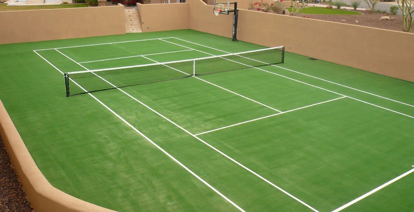 A professional sports field in Buckeye, Arizona, featuring a well-maintained green tennis court surrounded by beige walls. The net is set up in the middle, and a basketball hoop is attached to one of the walls. Stairs leading up are visible in the background. Red and green plants border the exterior areas.