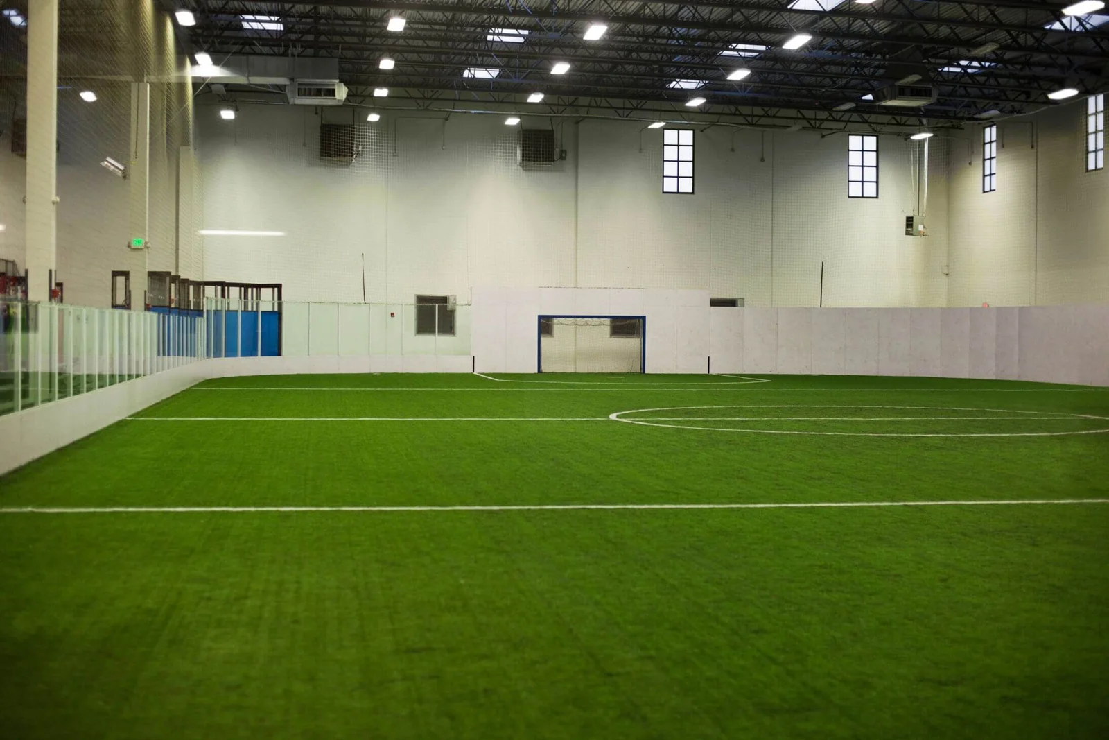 An indoor soccer field in Buckeye, Arizona, featuring artificial grass outlined with white lines. The field is surrounded by transparent barriers on two sides and white walls at the far end. Overhead, large lights illuminate the space, while windows line the upper part of the walls.