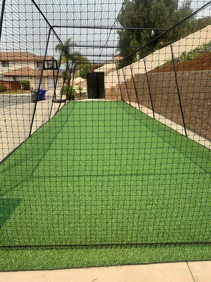 A long rectangular batting cage with green artificial turf ground is enclosed by netting. The cage, reminiscent of a sports field, is set up in a Buckeye, Arizona backyard with a brick wall along the side and houses visible in the background. There is a basketball hoop and trash bins on the left side.