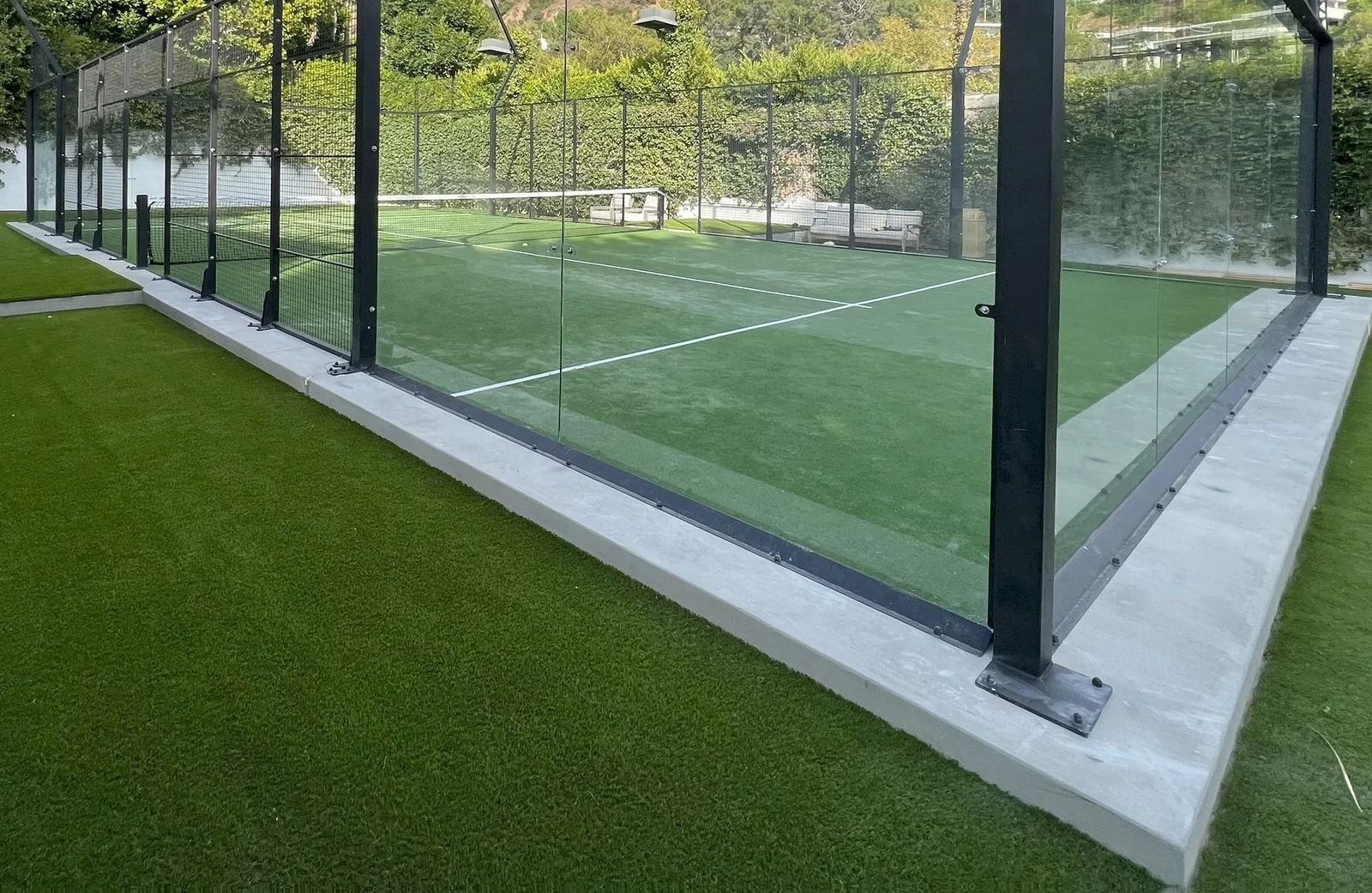 A modern, outdoor paddle tennis court in Buckeye, Arizona, enclosed with transparent glass walls and surrounded by lush green artificial turf. The court has a net, white playing lines, and is set against a backdrop of dense foliage and trees, showcasing top-tier artificial grass installations.