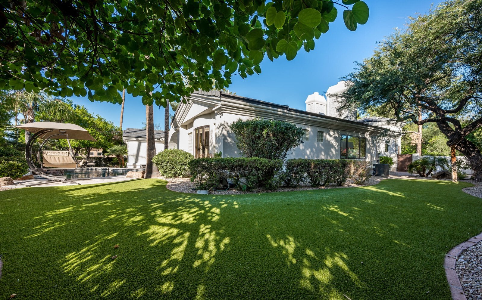 A modern house with a well-maintained backyard featuring lush green synthetic grass, manicured shrubs, and shaded areas from surrounding trees. In the background, there's a pool area with a cushioned swing and patio furniture under a partially shaded canopy. The sky is clear and blue.