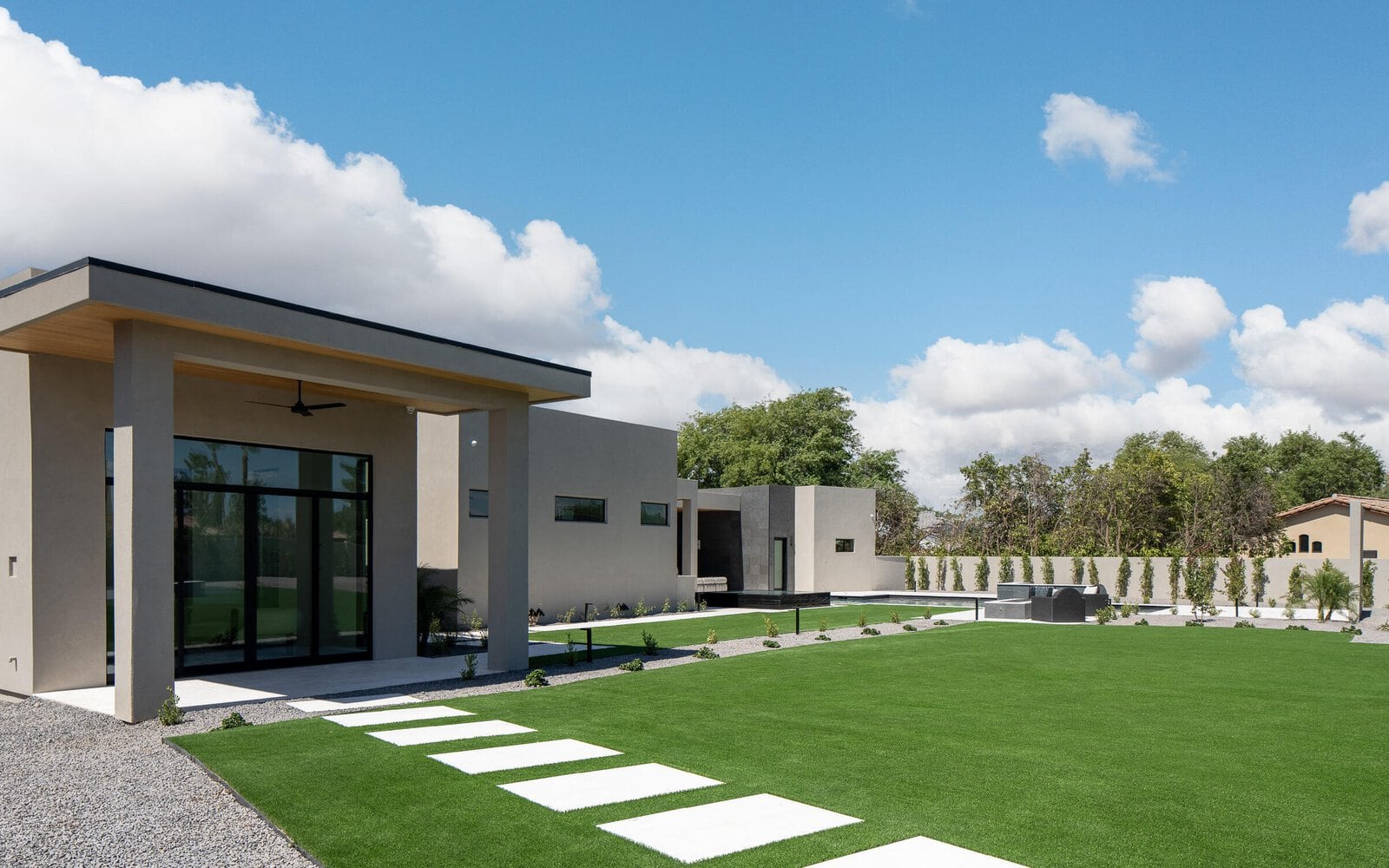 A modern house with a minimalistic design in Buckeye, AZ features large glass doors and windows, a flat roof, and beige exterior walls. The front yard boasts a low-maintenance lawn expertly crafted by an artificial grass installer, with stepping stones leading to the entrance. Trees and a clear blue sky with clouds are in the background.