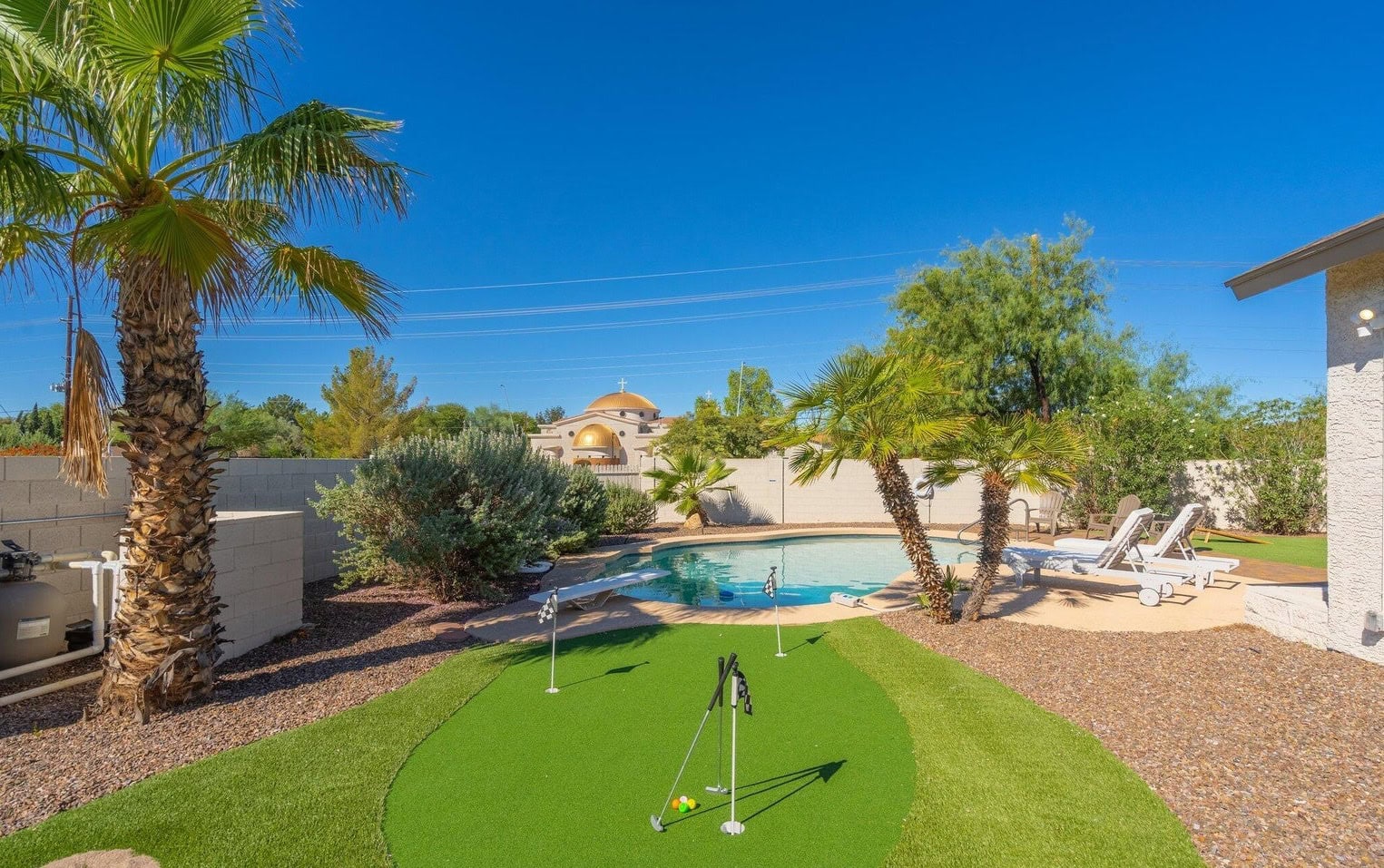 A backyard features a mini putting green with low-maintenance turf and a swimming pool surrounded by palm trees and lush greenery. Lounge chairs sit on the pool deck, and a stone wall encloses the space. The sky is clear and blue, suggesting a sunny day.