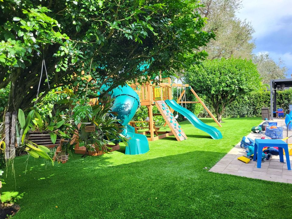 A lush backyard in Buckeye, AZ features a wooden playset with green and blue slides, climbing walls, and a swing. The playset is shaded by trees, surrounded by low-maintenance lawns. A blue table and scattered toys rest on a stone patio to the right, under a cloudy sky.